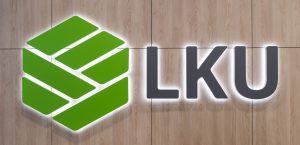 LKU logotipas ant medienos plokštės sienos - žalias logotipo elementas ir raidės "LKU"